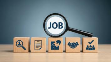 Image representing recruitment and job opportunities. A magnifying glass highlighting the word “JOB” is positioned above a row of wooden blocks displaying icons for candidate search, a résumé, skills or problem-solving, partnership, and successful hiring.