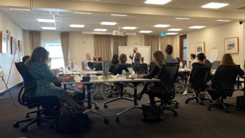 An image of a classroom with adult people at tables with a facilitator in front of a whiteboard