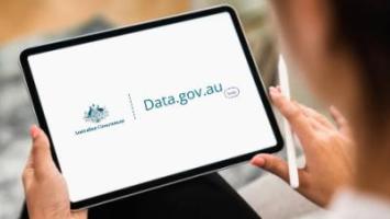 Person holding a tablet displaying the Data.gov.au website and Australian Government branding. The screen shows the Data.gov.au logo, while a stylus is held in the other hand, representing access to government data and digital information services.