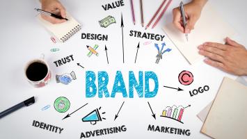 Brand strategy workshop graphic showing the word “BRAND” at the centre, connected to concepts including Value, Strategy, Design, Trust, Identity, Advertising, Marketing and Logo. Two people write in notebooks around the diagram, illustrating collaborative planning and brand development.