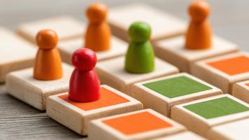 Colourful wooden game pieces placed on square tiles arranged in a grid. The pieces occupy different positions across the board, illustrating organisation, participation, resource allocation, team structures or strategic planning.